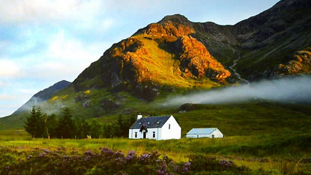 Photo of a small white cottage and outbuilding located on a green field with a small mountain behind.