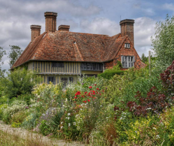 A Tudor style house with a tall gable roof and four prominent chimneys, set back behind a lush English garden of flowers, trees, and shrubs.